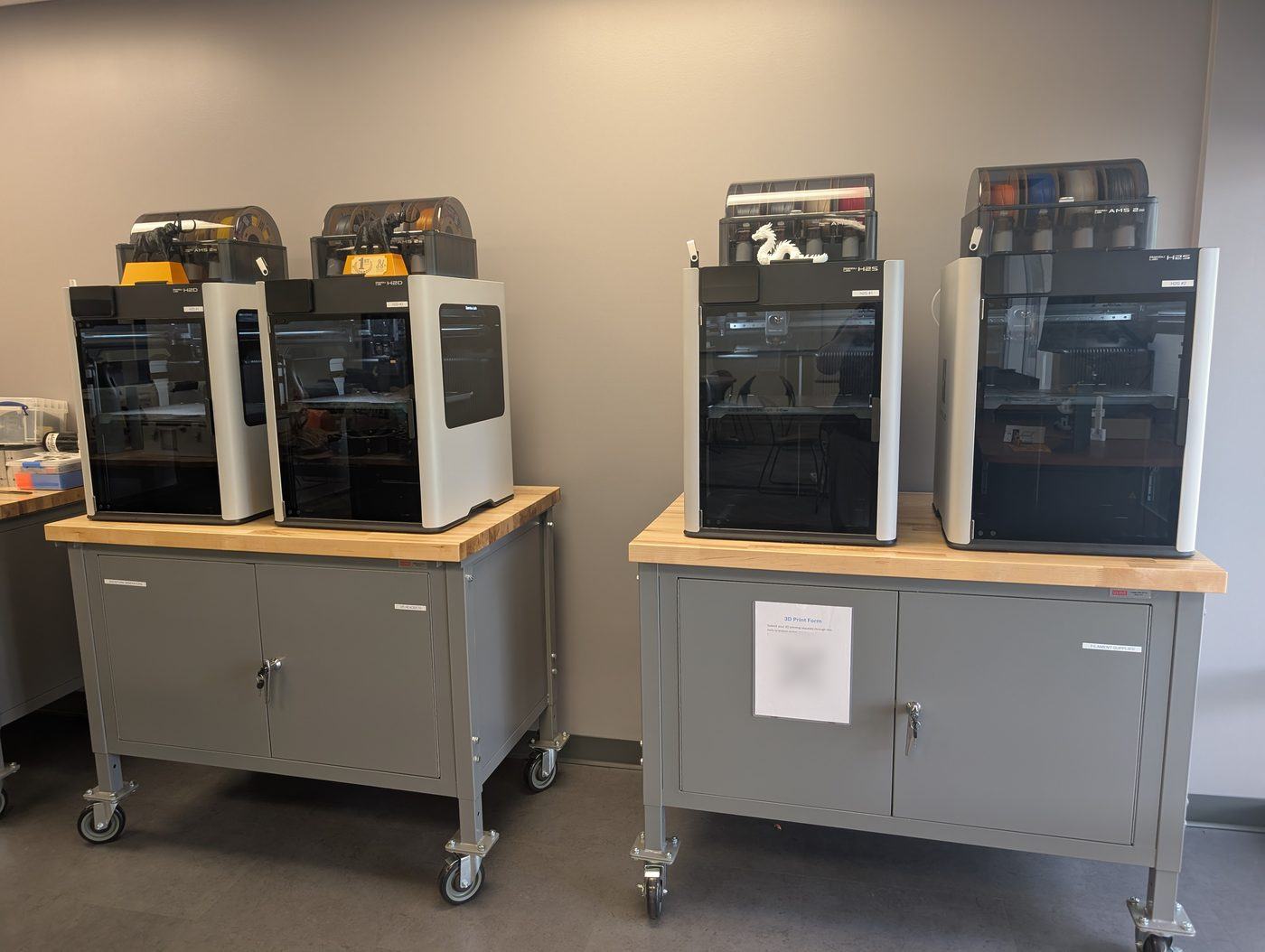 Four enclosed 3D printers on two rolling workbenches, with spools of filament on top.