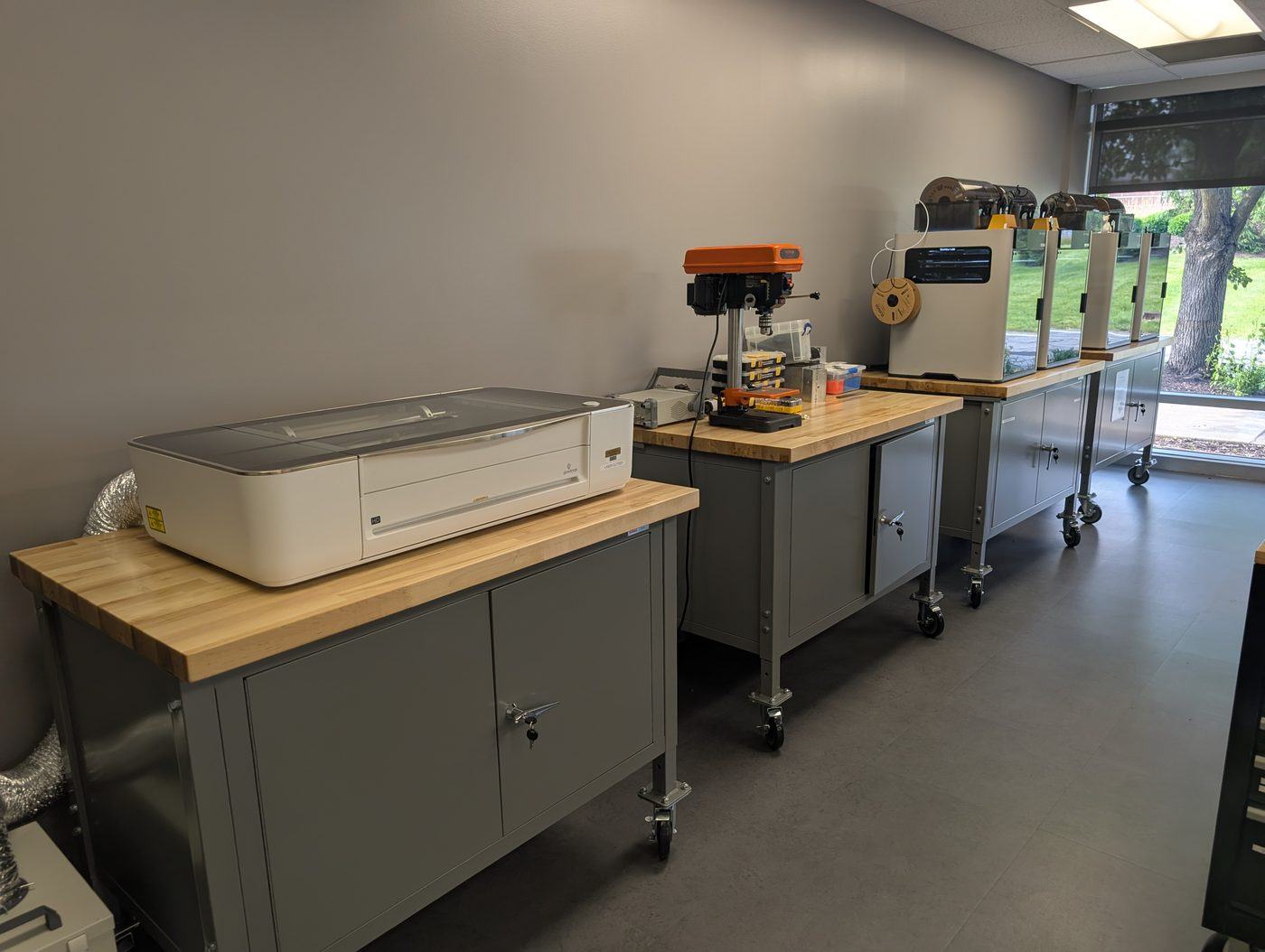 A white desktop laser cutter on a workbench, with a drill press and 3D printers on the benches beyond it.
