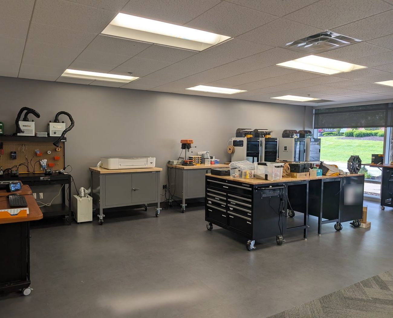 The Innovation Lab: an open room with rolling workbenches, a soldering station with fume extractors, a laser cutter, a drill press and a row of 3D printers beside a large window.