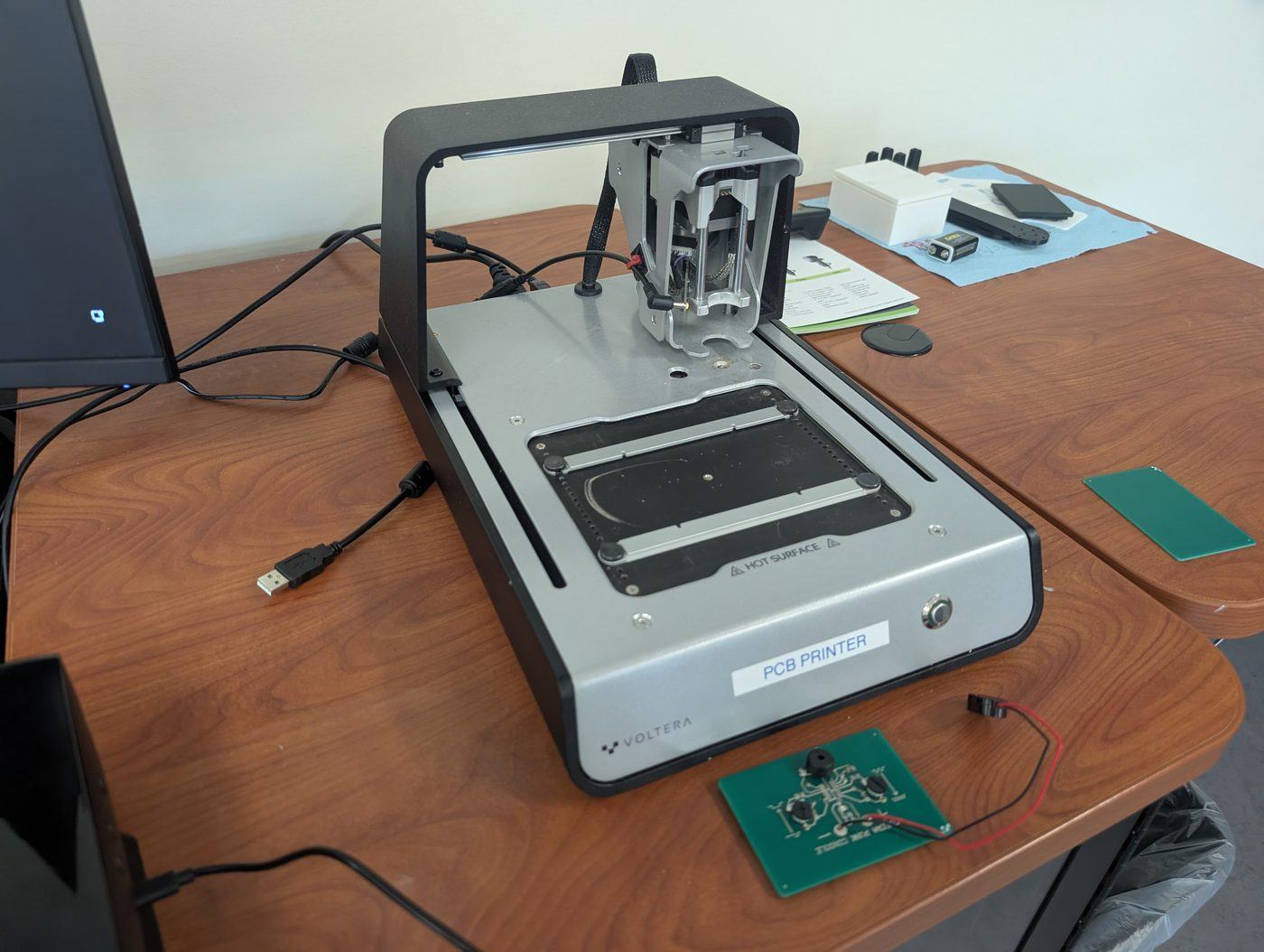 A desktop circuit-board printer on a table, with a finished green circuit board in front of it.